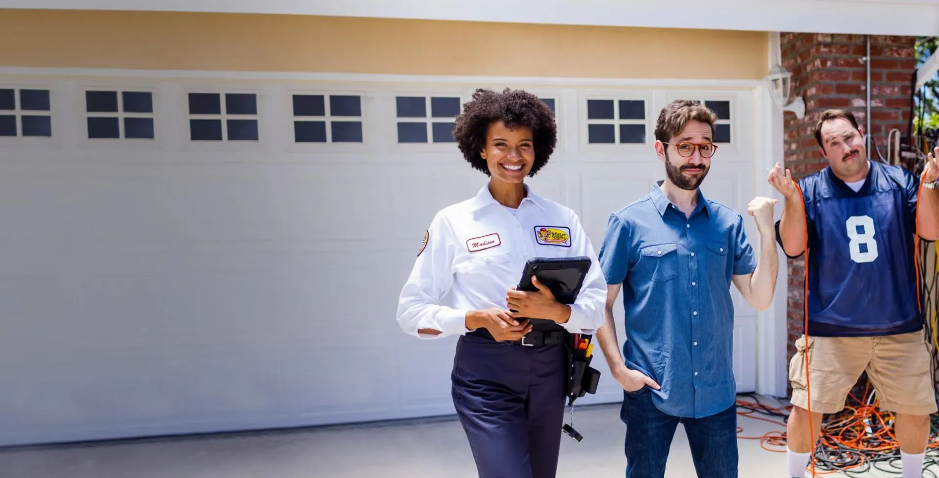 Female electrician with a tablet smiling at the camera next to two customers in front of a garage.