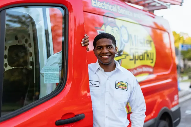 Female electrician with a tablet smiling at the camera next to two customers in front of a garage.